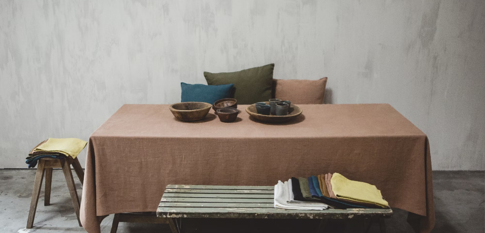 Libeco Handcrafted Collection featuring a brown tablecloth-covered table with wooden bowls, ceramic cups, and a plate, surrounded by pillows and folded fabrics in various colors
