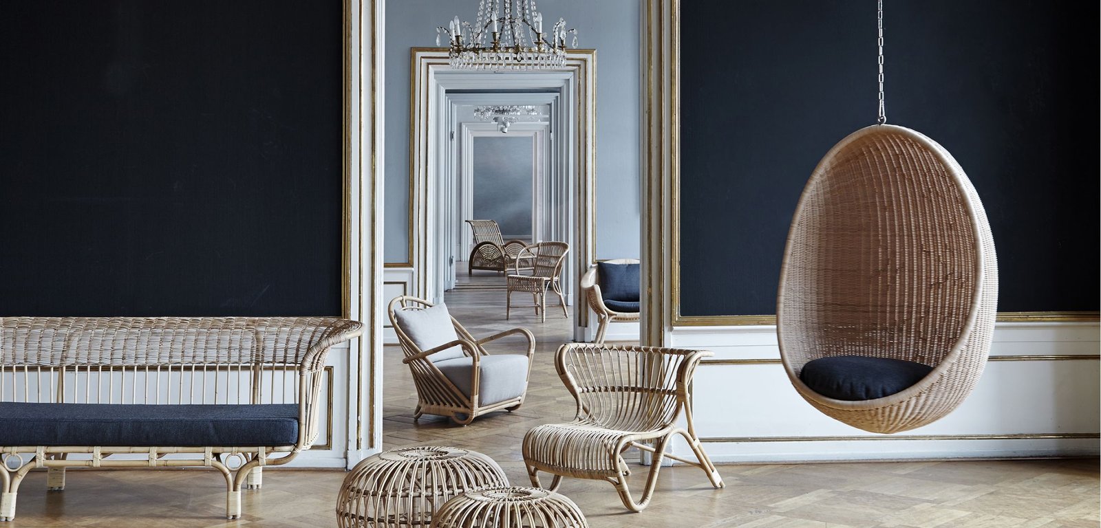 A collection of wicker furniture including a hanging chair, a bench, two chairs, and two ottomans, set against a dark wall with large ornate mirrors or doorways