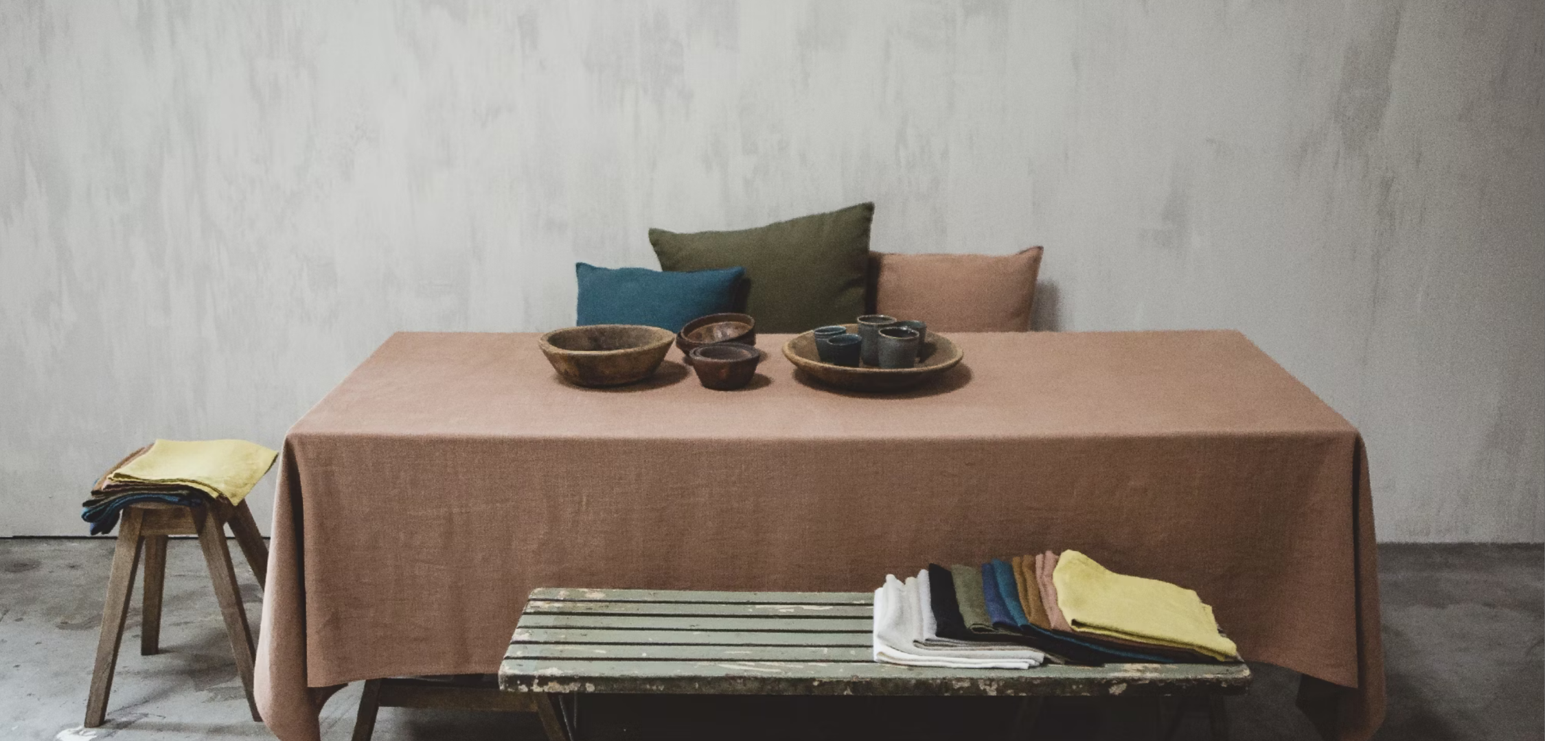 Libeco Handcrafted Collection featuring a brown tablecloth-covered table with wooden bowls, ceramic cups, and a plate, surrounded by pillows and folded fabrics in various colors